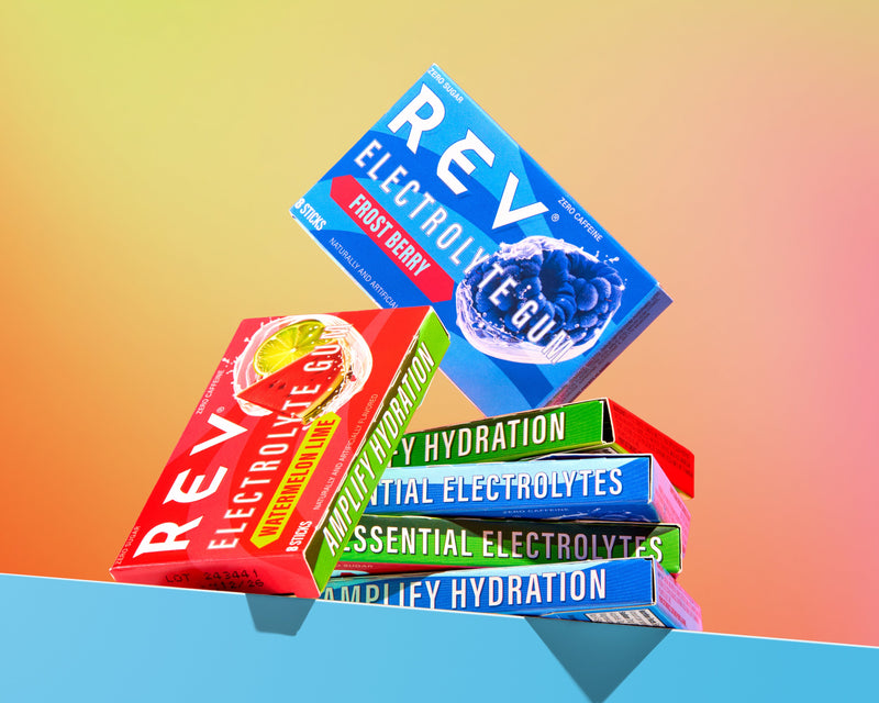 pile of rev watermelon lime electrolyte and rev frost berry electrolyte against a rainbow ombre background on top of a blue edge