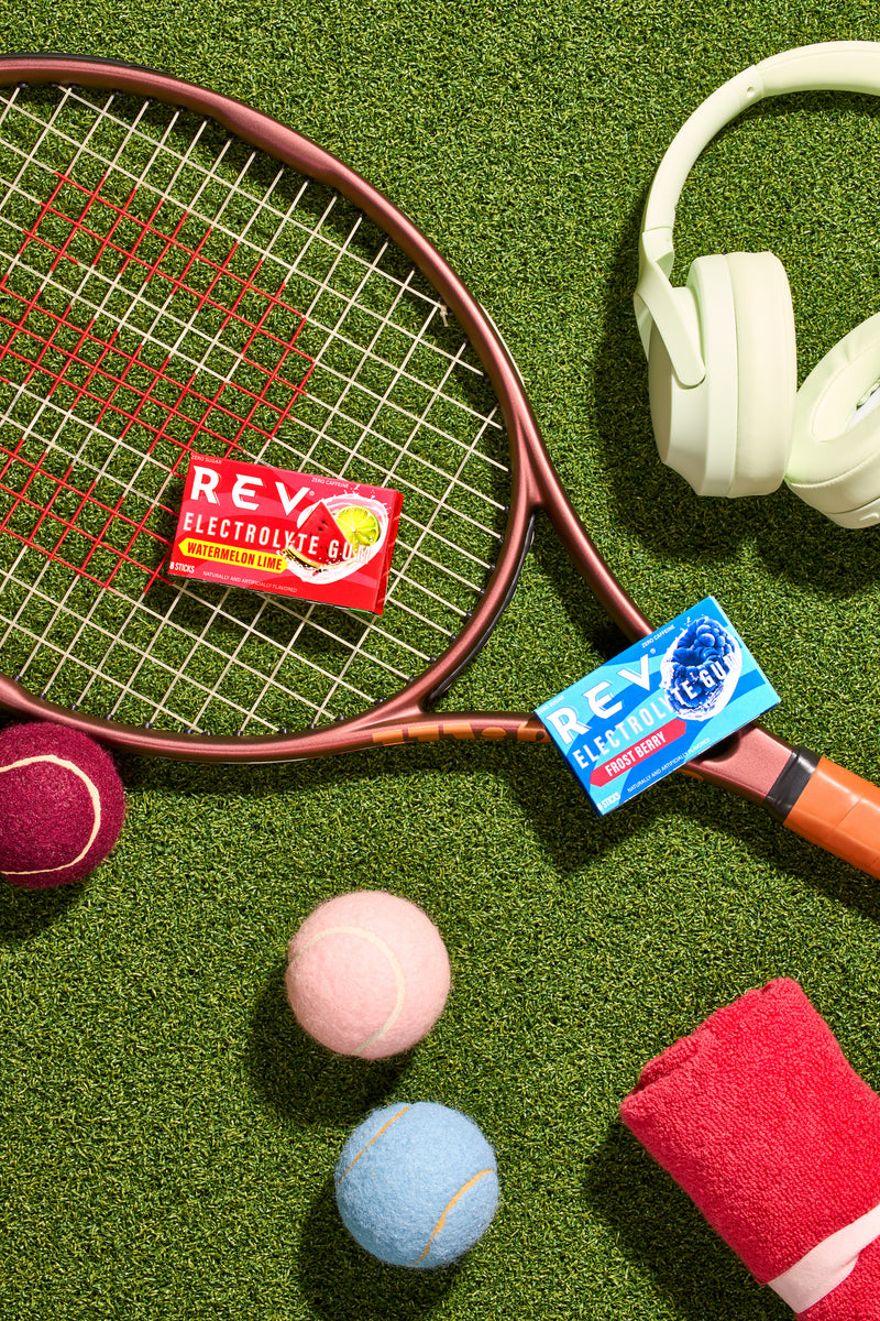 rev watermelon lime electrolyte gum and rev frost berry electrolyte gum on a tennis racket on turf background with tennis balls and headphones